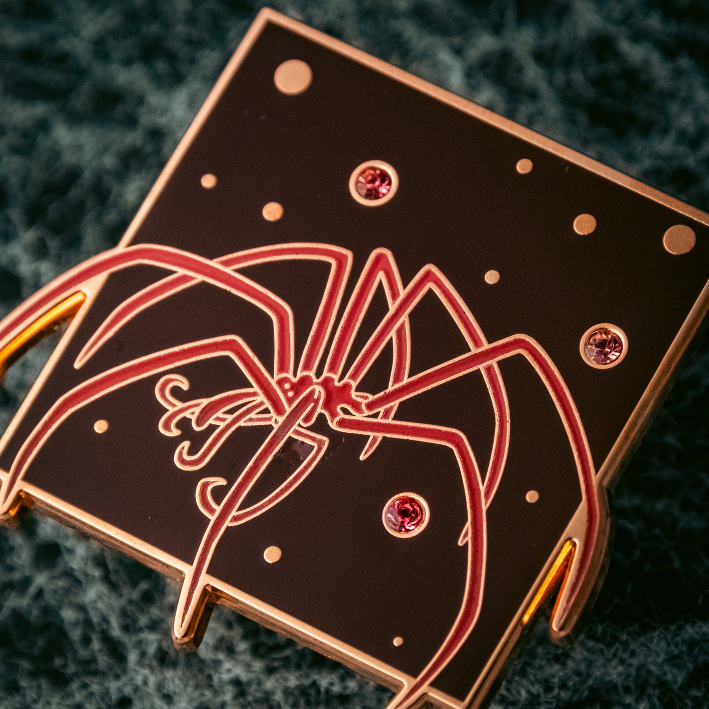 A photo of a soft enamel pin featuring a Sea Spider with red enamel coloring and copper plating with red gemstone inlays.