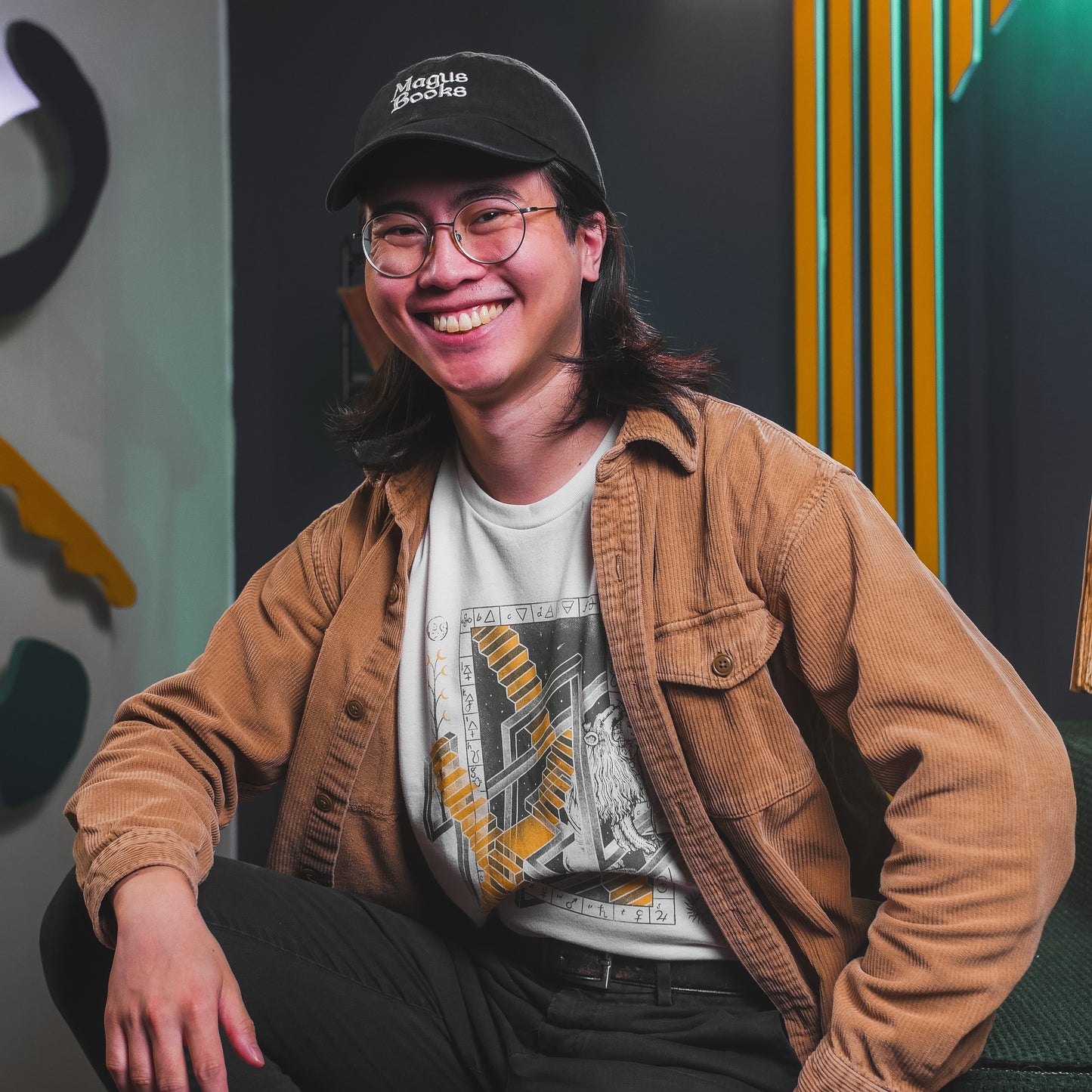 Tom Lum wearing an off-white t-shirt with a graphic in green and yellow featuring various alchemical symbols while sitting in the SciShow studio.