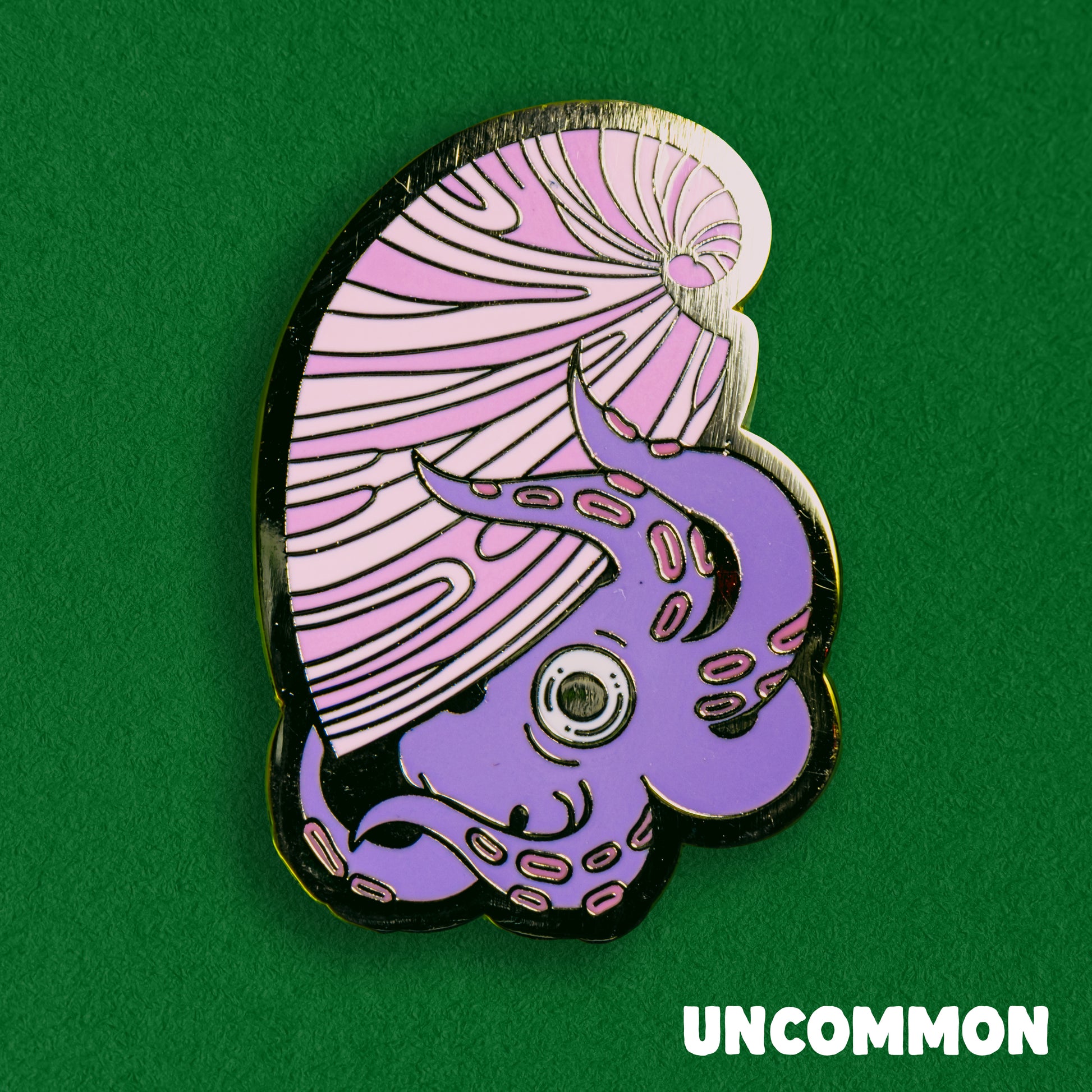 A photo of a soft enamel pin featuring an argonaut on a green background. It is purple and pink with gold metal plating. Uncommon