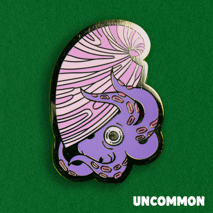 A photo of a soft enamel pin featuring an argonaut on a green background. It is purple and pink with gold metal plating. Uncommon