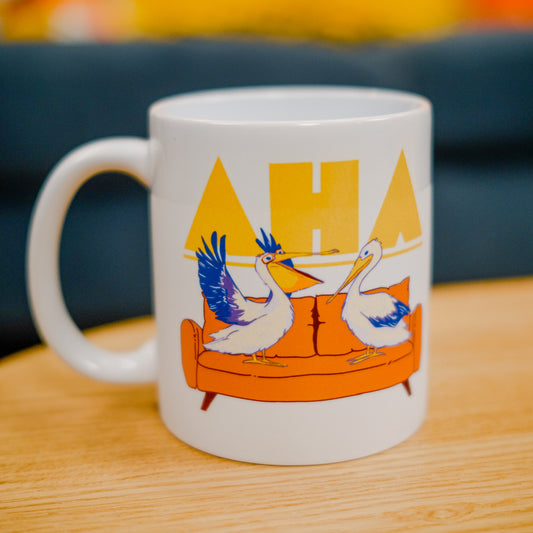 A photo of a white mug featuring a design of two pelicans on a couch. One wears glasses and has its wings spread as if gesturing. The Ask Hank Anything logo is behind them.