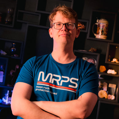 Hank Green wearing a blue t-shirt with a text-based design on the chest while standing in his office. The design reads "MRPS" in big text and "moon rock protection squad" in smaller text underneath it. From SciShow.