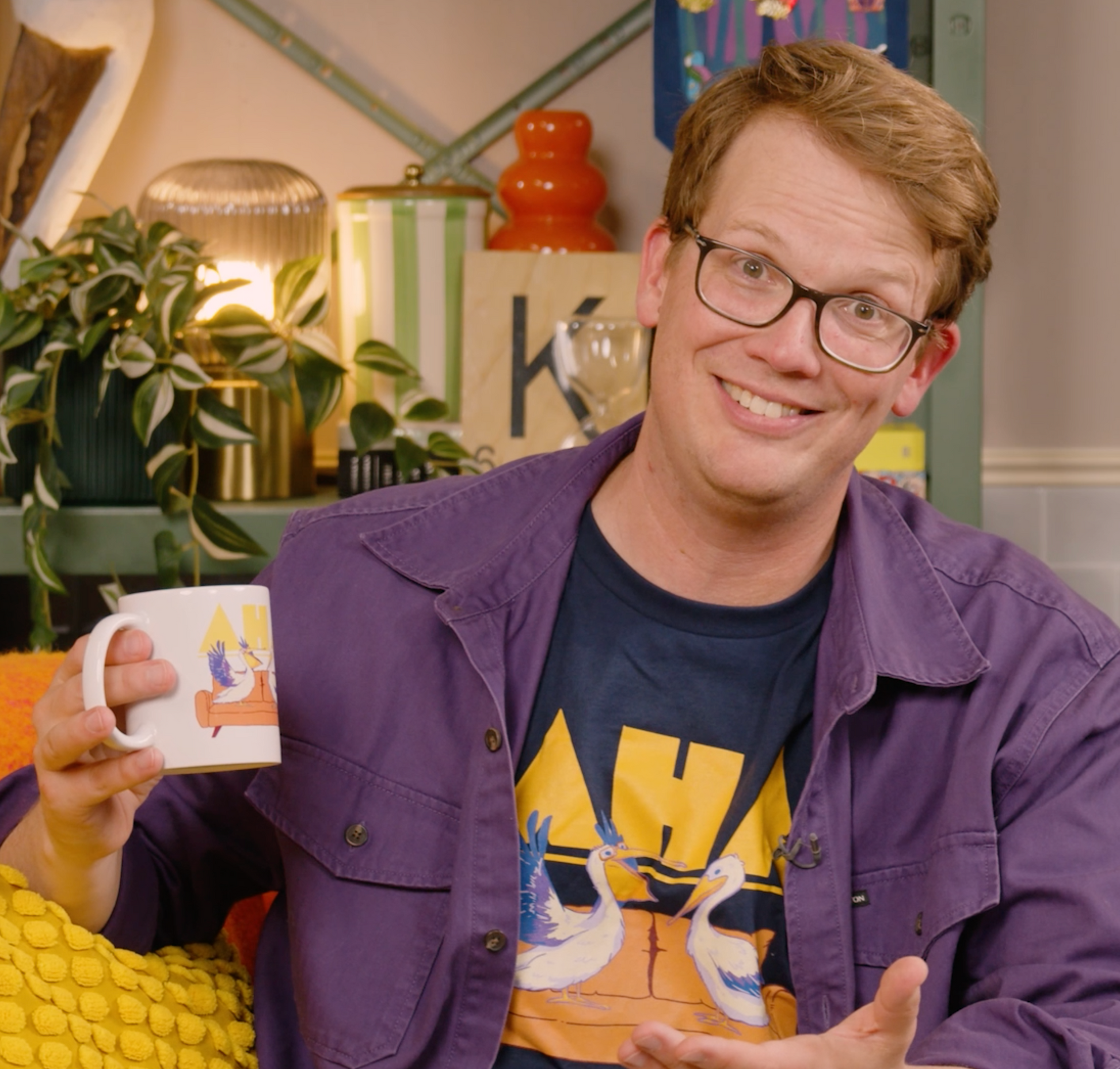 A photo of Hank Green holding the AHA Pelican mug while gesturing. He wears the AHA Pelican shirt, which is navy with the same design.