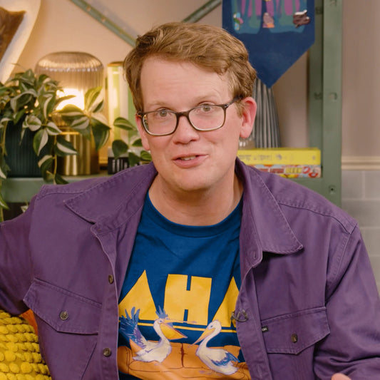 A photo of Hank Green wearing the AHA Pelican shirt underneath a purple shirt jacket.