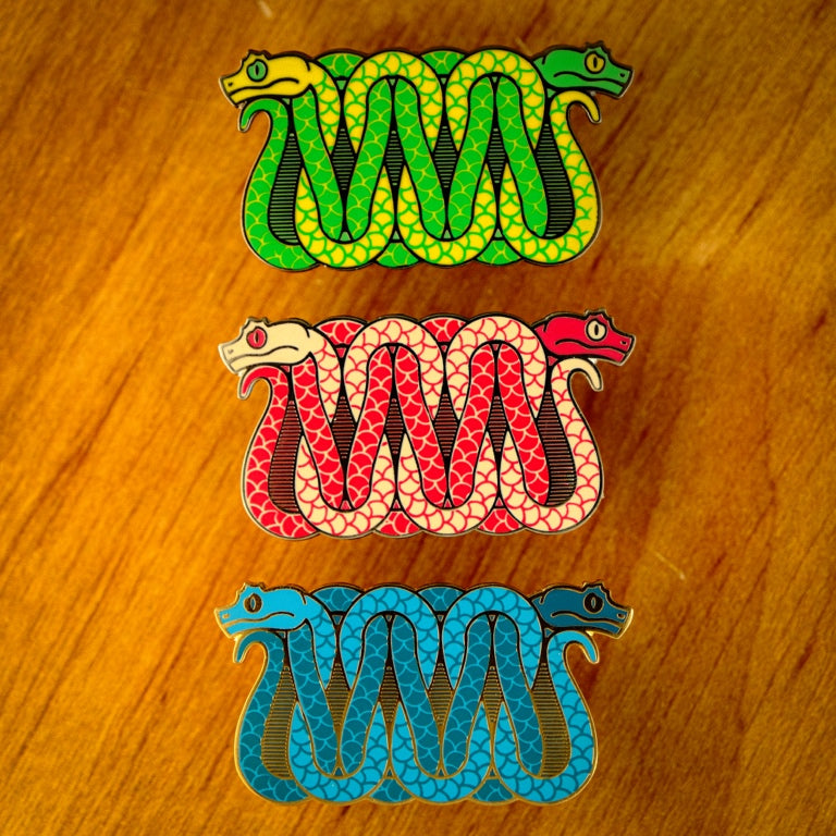 A photo of 3 soft enamel pins featuring eyelash vipers. The first is blue with green and yellow with black metal, the second is cream and red with black metal, and the third is blue and teal with gold metal. 