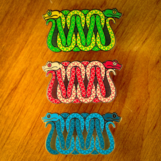 A photo of 3 soft enamel pins featuring eyelash vipers. The first is blue with green and yellow with black metal, the second is cream and red with black metal, and the third is blue and teal with gold metal. 
