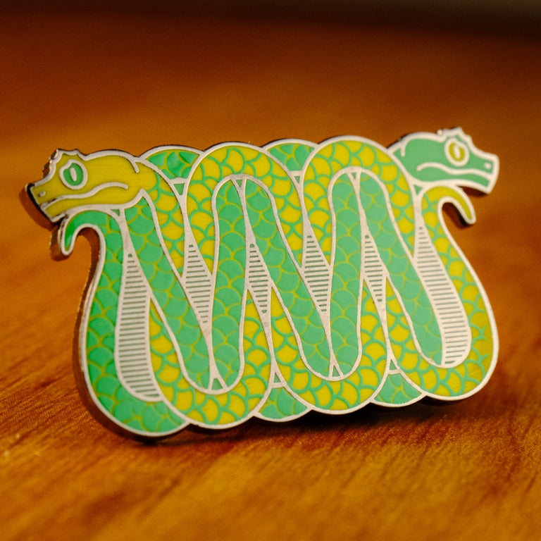 A photo of a soft enamel pin featuring eyelash vipers in green and yellow enamel with black metal plating. Common