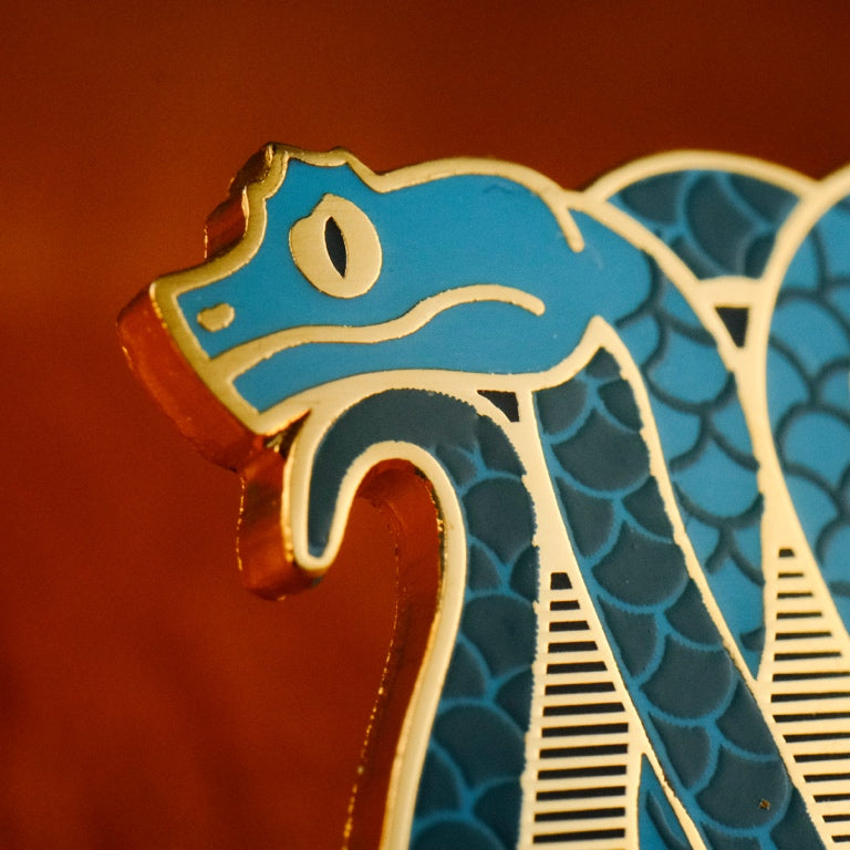A close up photo of a soft enamel pin featuring eyelash vipers in blue and teal enamel with gold metal plating, showcasing the metal plating as the eye with a black enamel fill for the iris.