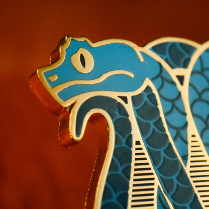 A close up photo of a soft enamel pin featuring eyelash vipers in blue and teal enamel with gold metal plating, showcasing the metal plating as the eye with a black enamel fill for the iris.