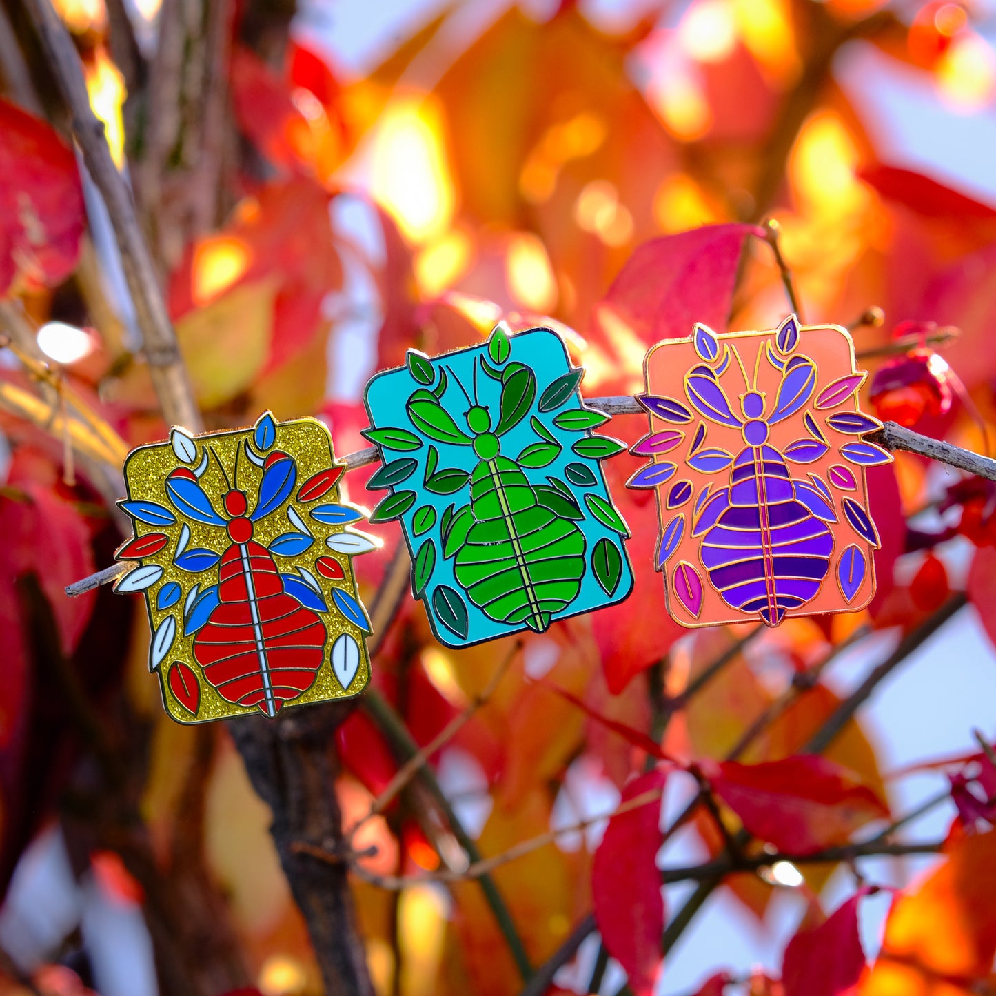 A photo of 3 soft enamel pins featuring Phasmids on a leafy autumnal background. The first is red and blue with gold enamel on a gold glittery background, the second is green with silver metal on a blue background, and the third is purple and pink with gold metal on a peach background. 