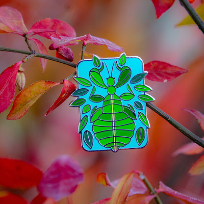 A photo of a soft enamel pin featuring a phasmid on a leafy autumnal background. It is green with silver metal on a blue background. Common