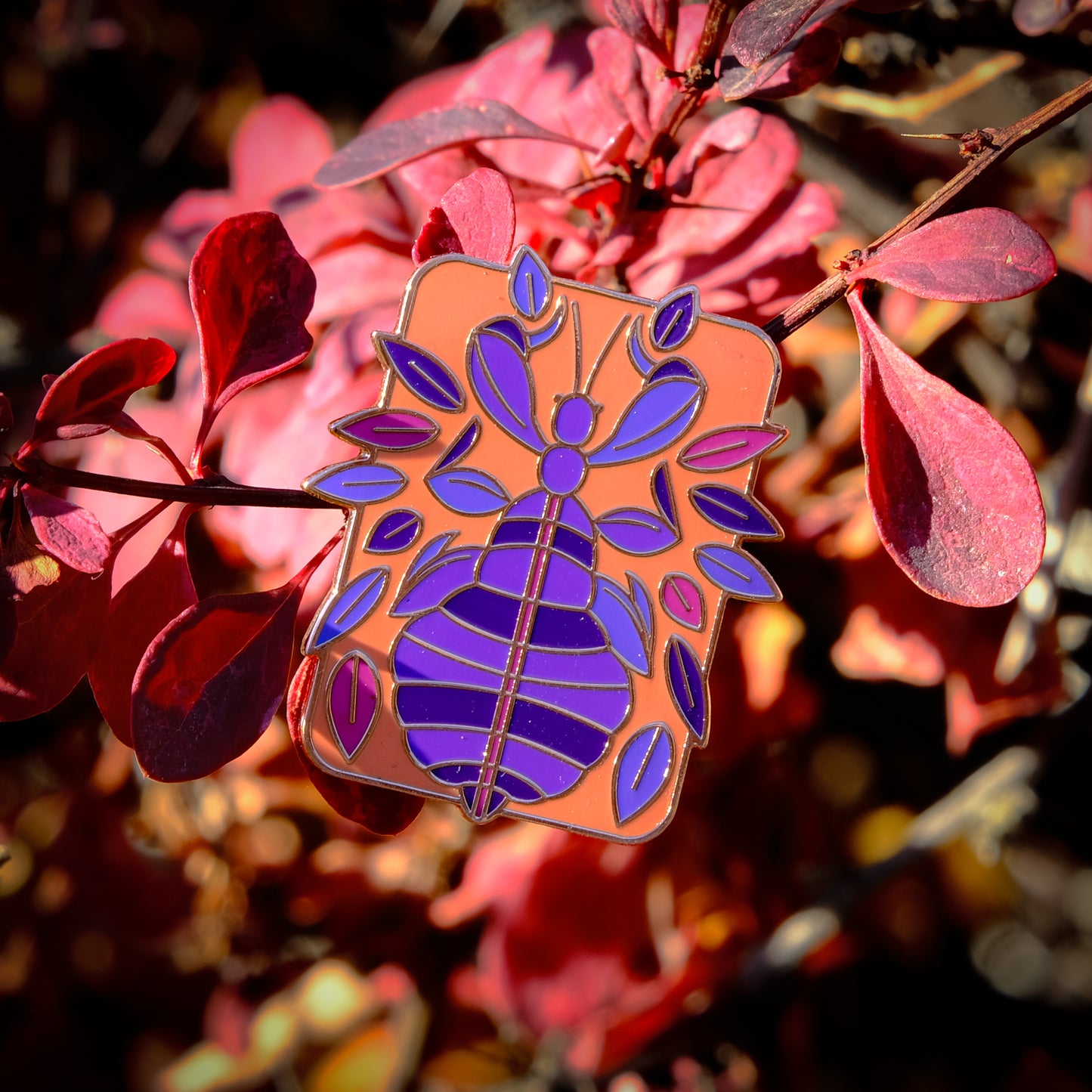 A photo of a soft enamel pin featuring a phasmid on a leafy autumnal background. It is pink and purple with gold metal on a peach background. Uncommon