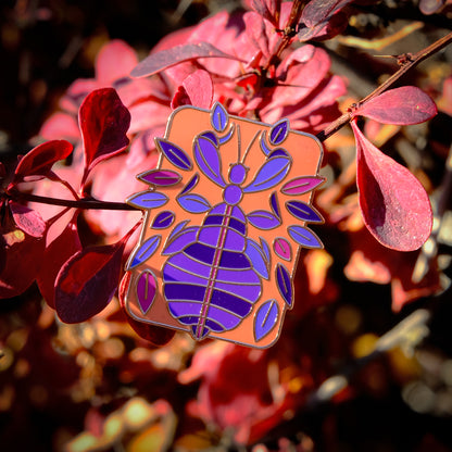 A photo of a soft enamel pin featuring a phasmid on a leafy autumnal background. It is pink and purple with gold metal on a peach background. Uncommon