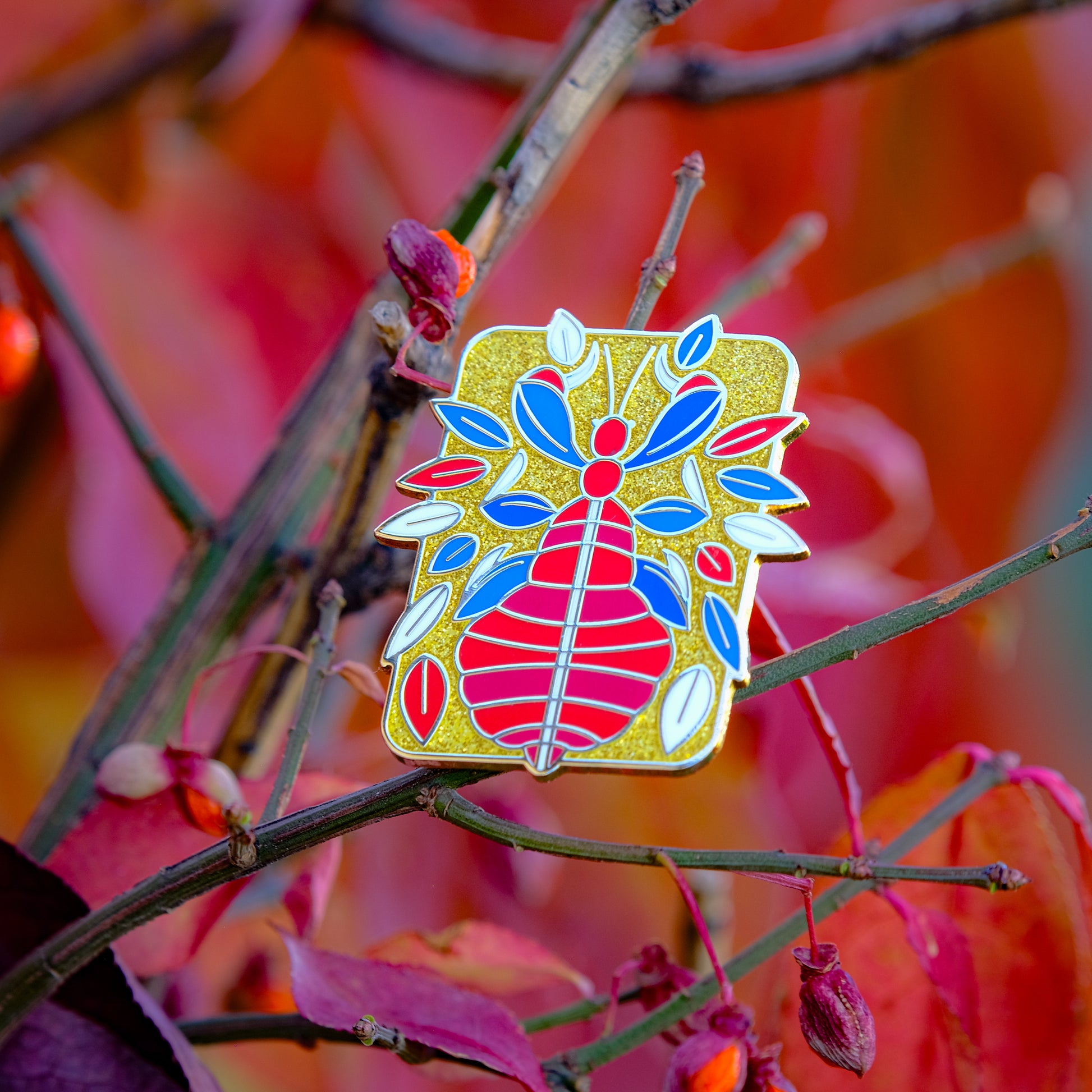 A photo of a soft enamel pin featuring a phasmid on a leafy autumnal background. It is red and blue with gold metal on a glittery gold background. Rare