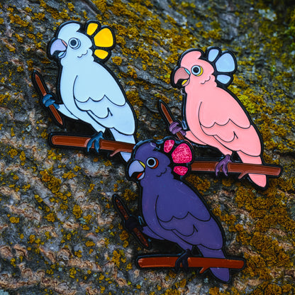 A photo of 3 soft enamel pins featuring Goffin's Cockatoos on a wood bark background. They all have black metal plating. The first has white and yellow enamel, the second has pink and lavender enamel, and the third has purple and pink enamel with glitter accents. 