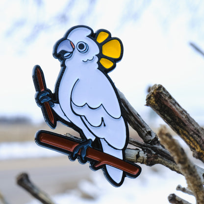A photo of a soft enamel pin featuring a Goffin's Cockatoo on a sparse wooded background. It is black-metal plated with white and yellow enamel. Common