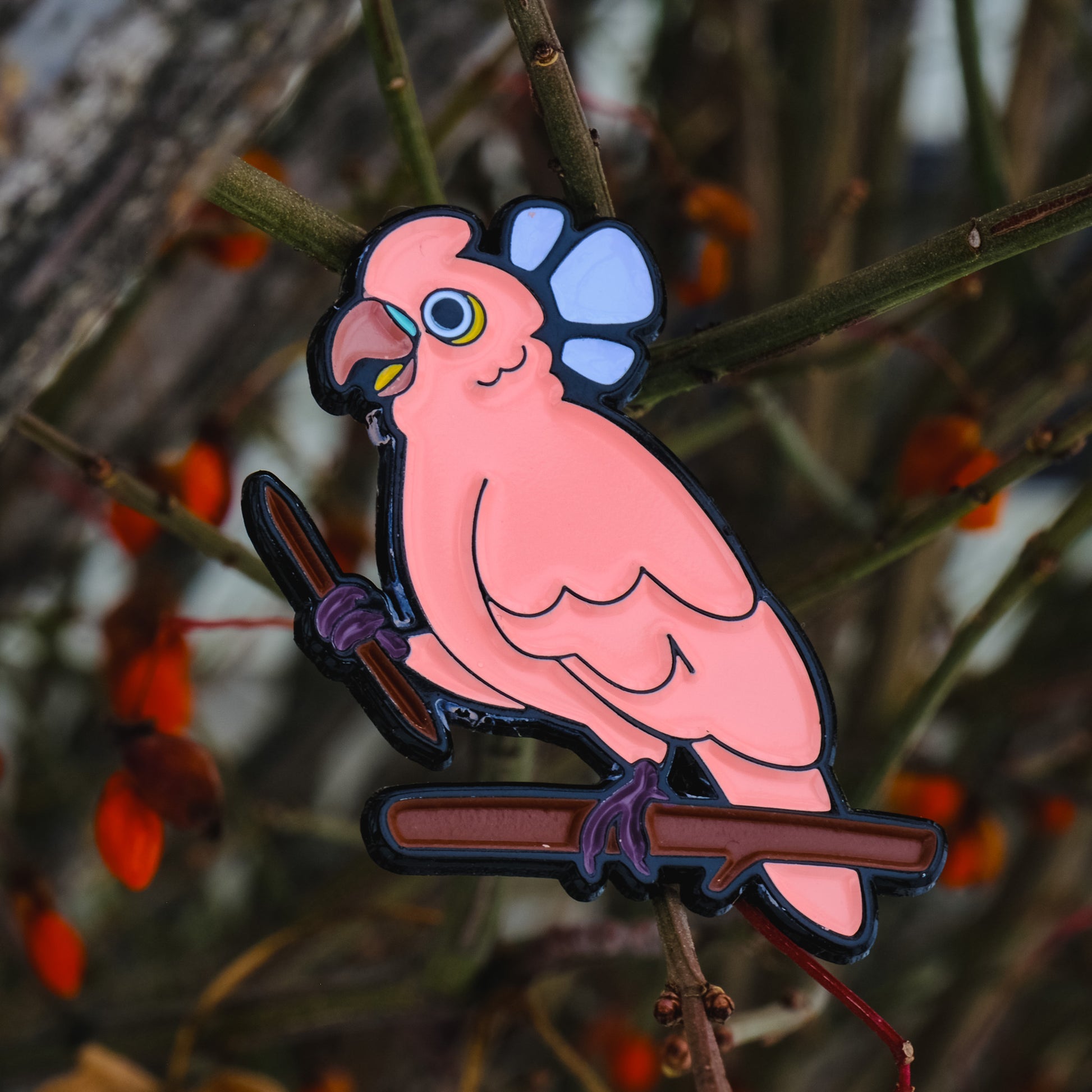 A photo of a soft enamel pin featuring a Goffin's Cockatoo on a sparse wooded background. It is black-metal plated with pink and lavender enamel. Uncommon