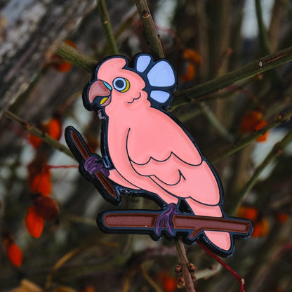 A photo of a soft enamel pin featuring a Goffin's Cockatoo on a sparse wooded background. It is black-metal plated with pink and lavender enamel. Uncommon