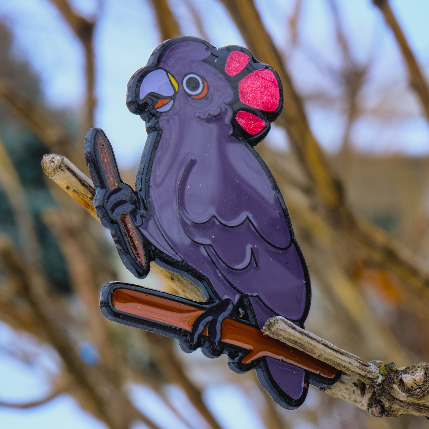A photo of a soft enamel pin featuring a Goffin's Cockatoo on a sparse wooded background. It is black-metal plated with purple and pink enamel and glitter accents. Rare
