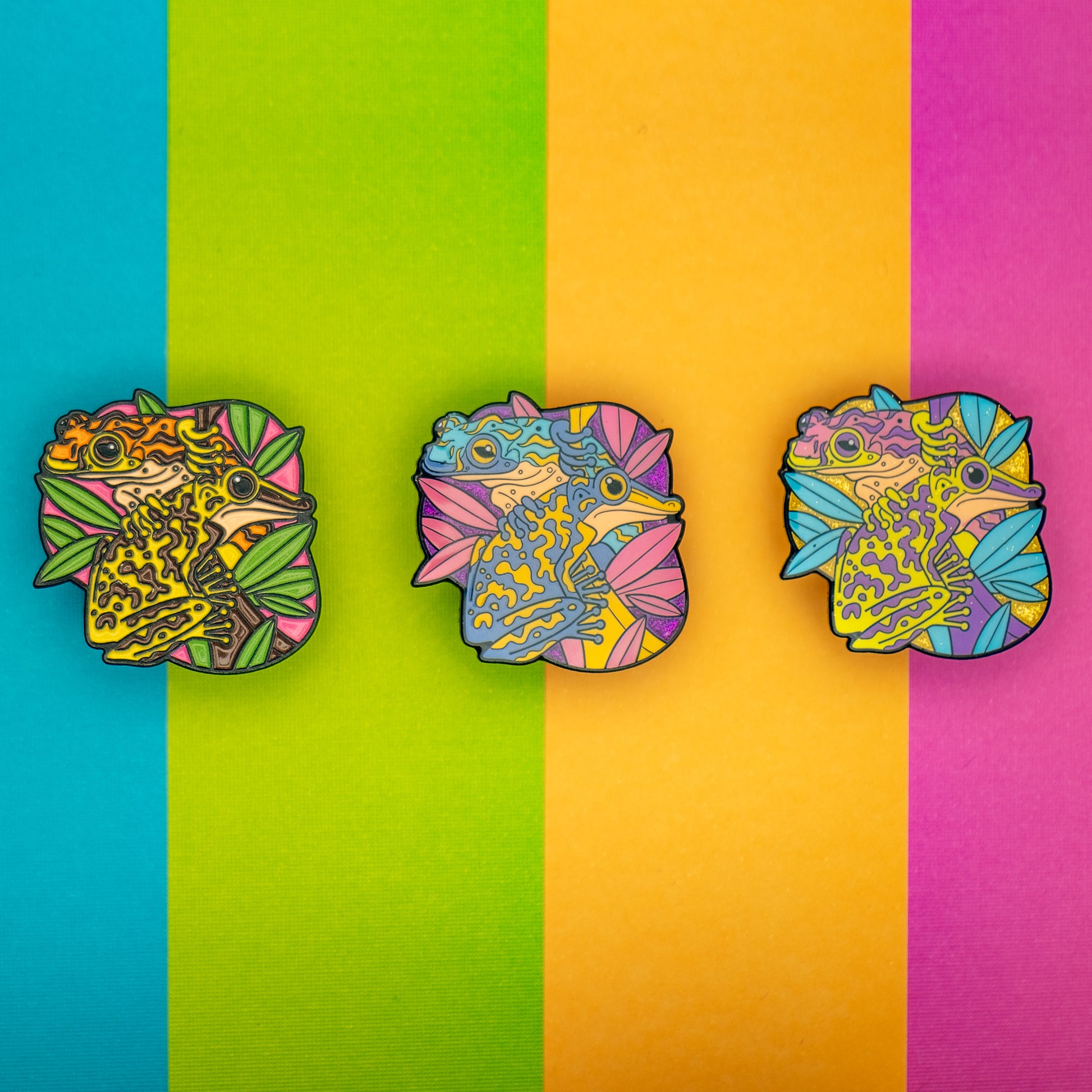 A photo of 3 soft enamel pins featuring Venomous Frogs on a colorful striped background. The first has yellow and brown frogs, the second has yellow and blue frogs with a glittery purple background, and the third has green and purple frogs with a glittery background. 