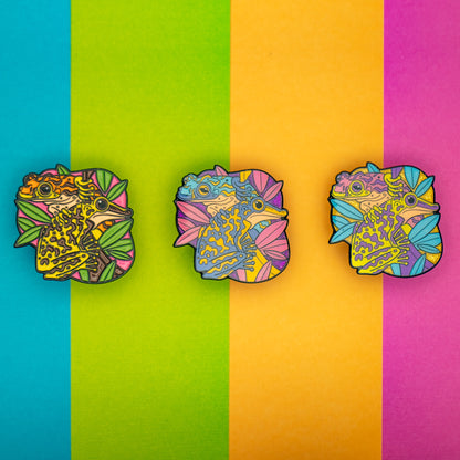 A photo of 3 soft enamel pins featuring Venomous Frogs on a colorful striped background. The first has yellow and brown frogs, the second has yellow and blue frogs with a glittery purple background, and the third has green and purple frogs with a glittery background. 