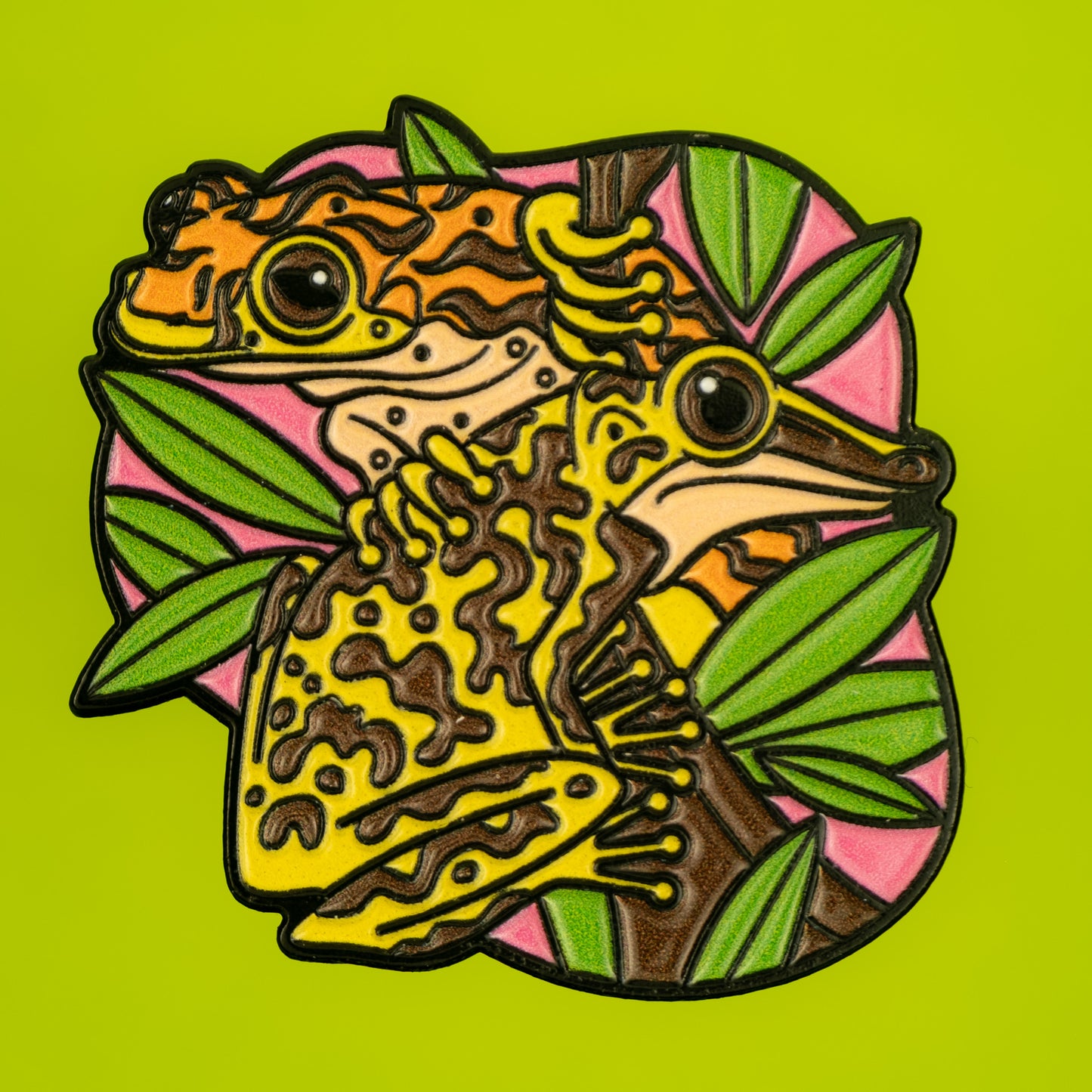 A photo of a soft enamel pin featuring Venomous Frogs on a bright green background. It has yellow and brown frogs. Common