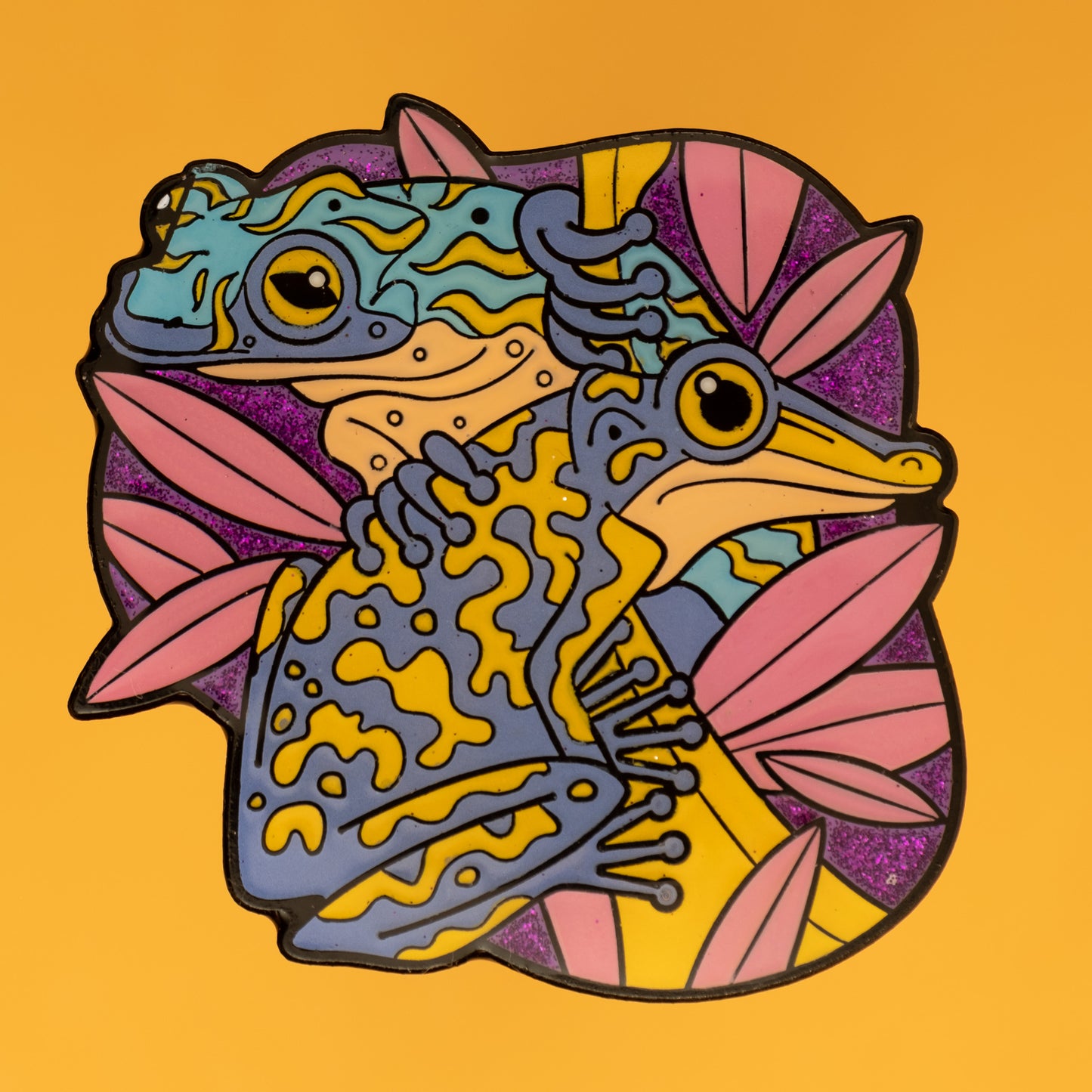 A photo of a soft enamel pin featuring Venomous Frogs on a light orange background. It has yellow and blue frogs with a glittery purple background. Uncommon
