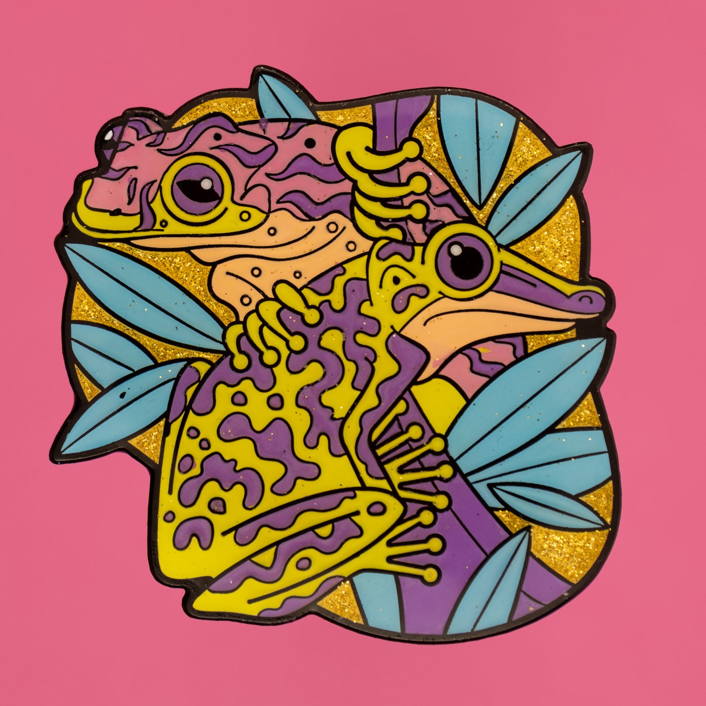 A photo of a soft enamel pin featuring Venomous Frogs on a bright pink background. It has green and purple frogs with a glittery yellow background. Rare