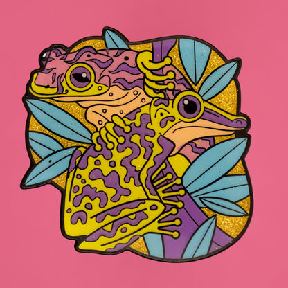 A photo of a soft enamel pin featuring Venomous Frogs on a bright pink background. It has green and purple frogs with a glittery yellow background. Rare
