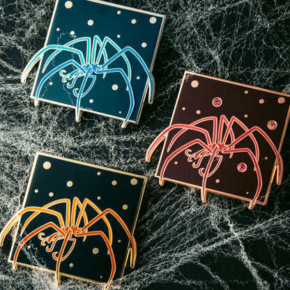 A photo of 3 soft enamel pins featuring Sea Spiders. The first is blue with silver plating, the second is red with copper plating and gem inlays, and the third is orange with gold plating.