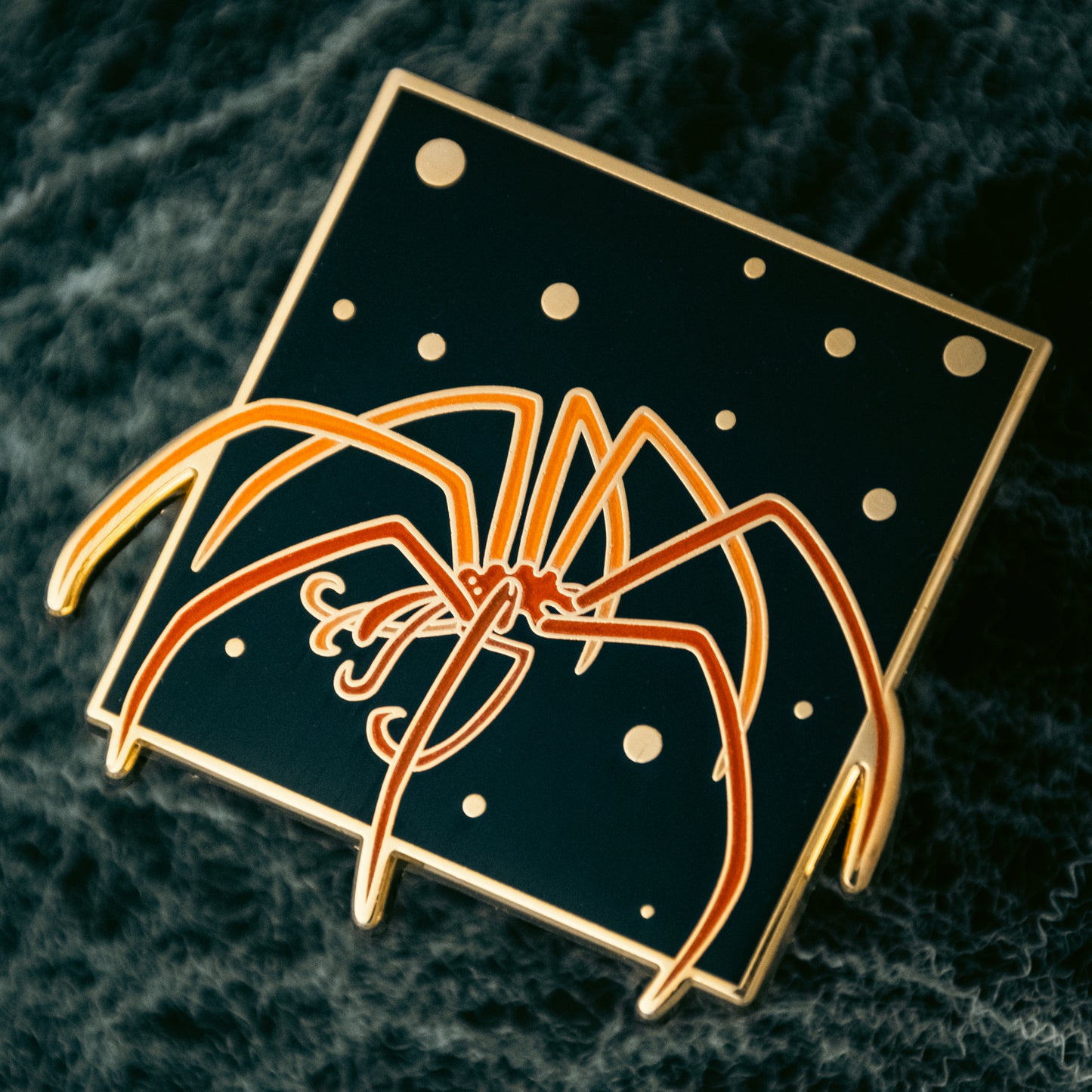 A photo of a soft enamel pin featuring a Sea Spider with orange enamel coloring and gold plating. Common