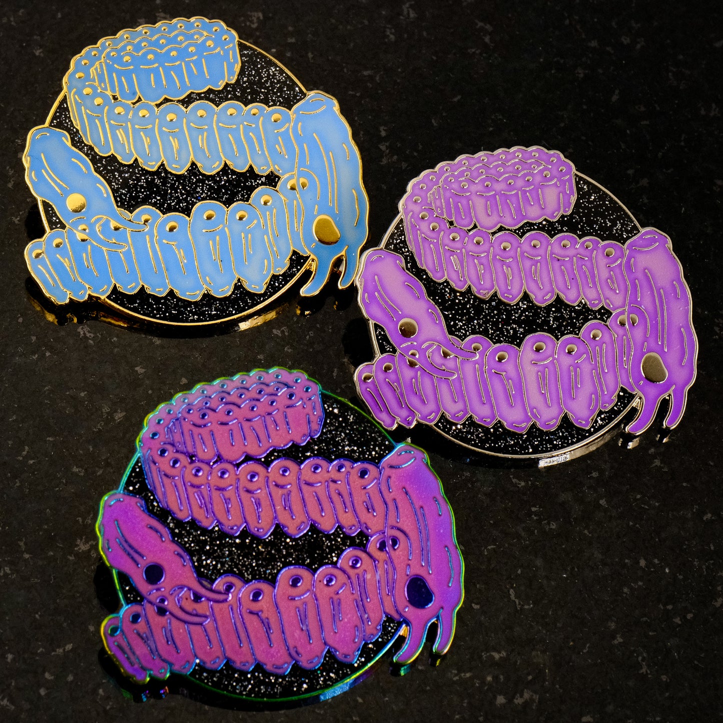 A photo of 3 soft enamel pins featuring Salps. The first is blue with gold metal, the second is light purple with gold metal, and the third is bright magenta with iridescent metal. All have glittery black backgrounds
