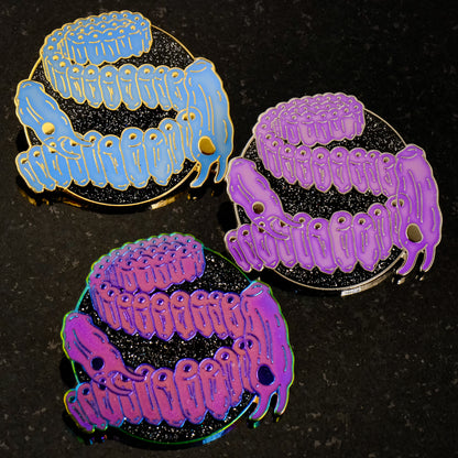 A photo of 3 soft enamel pins featuring Salps. The first is blue with gold metal, the second is light purple with gold metal, and the third is bright magenta with iridescent metal. All have glittery black backgrounds