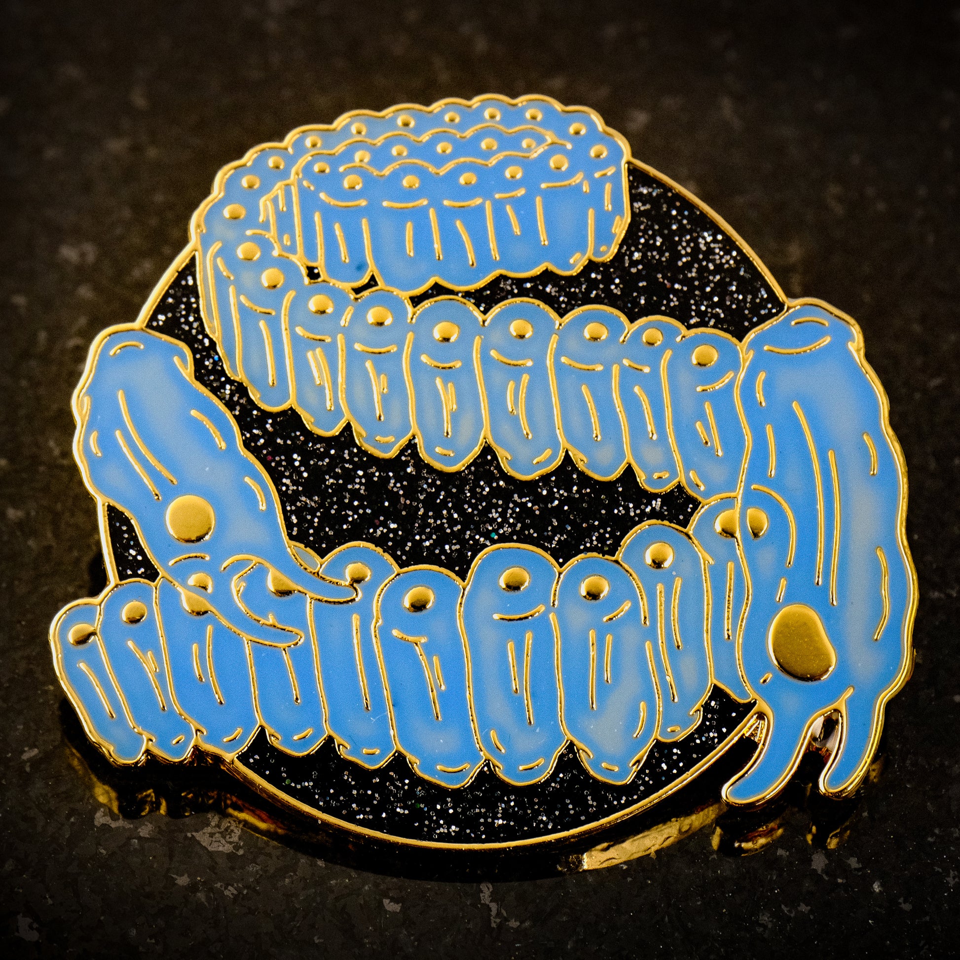 A photo of a soft enamel pin featuring Salps on a glittery black background with gold metal and blue enamel. Common